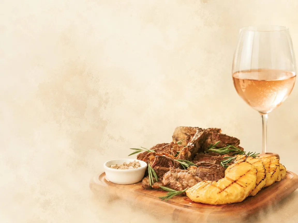 Board of grilled meat with roasted pineapple, rosemary and fleur de sel, and a glass of rosé wine, on a cream background